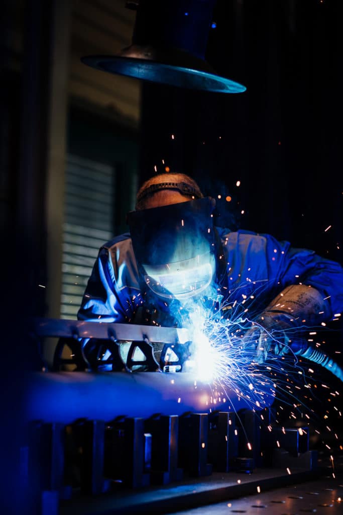 A person wearing a protective welding mask and gloves welds maschinenbauteile, creating bright blue sparks in a dark maschinenbau workshop.
