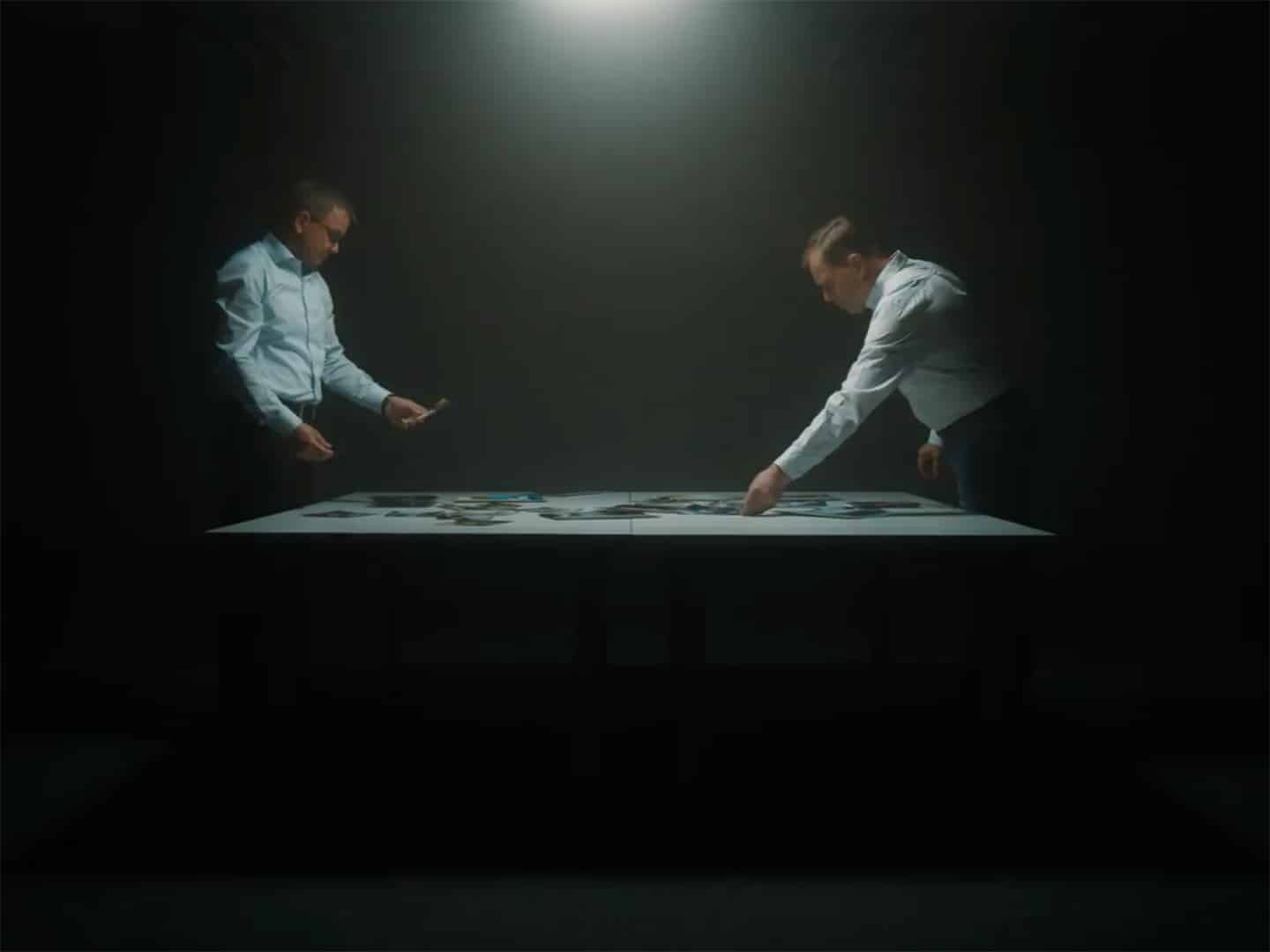 Zwei Männer in Oberhemden stehen sich an einem Tisch gegenüber und setzen im Scheinwerferlicht ein großes Puzzle zusammen. Die dramatische Szene, die an die Präzision von Kersten Maschinenbau erinnert, unterstreicht die konzentrierte Teamarbeit inmitten der schattigen Umgebung.